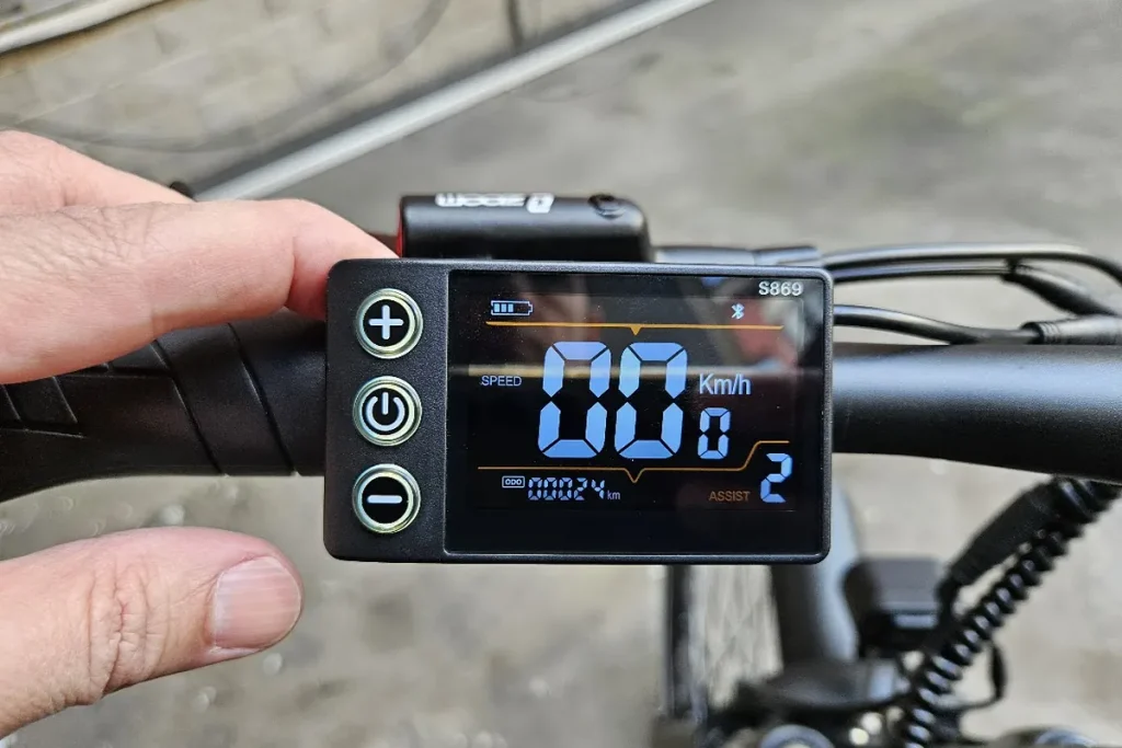 Écran S869 d'un vélo électrique et ses boutons de commande, main du cycliste sur la poignée.